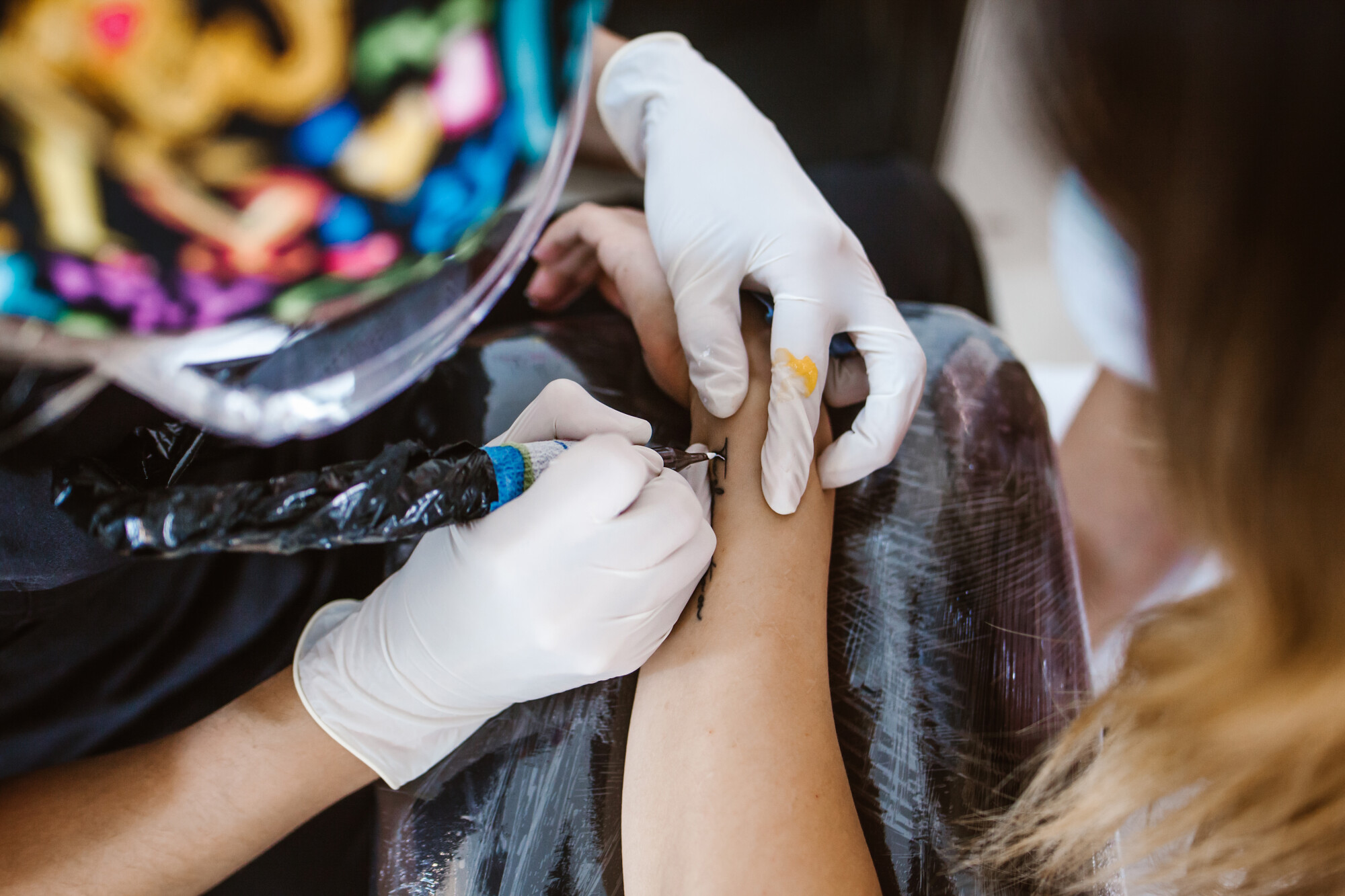 Woman being tattooed on the side of the wrist with a gloved person using tattoo equipment.